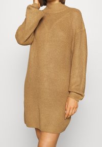 Stickad, långärmad klänning i ljusbrunt med hög hals och lös passform; har texturerad tyg och ribbade ärmändar.