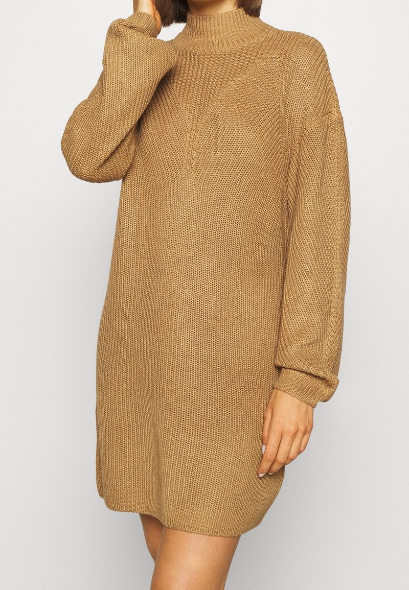 Stickad, långärmad klänning i ljusbrunt med hög hals och lös passform; har texturerad tyg och ribbade ärmändar.