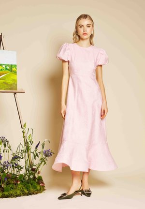 Jauna moteris vilki šviesiai rožinę midi su pūstomis rankovėmis suknelę ir žalius žemo kulno batelius su atvirais kulnais, stovinti prie molberto su peizažo tapyba.