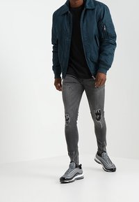 Chaqueta bomber verde azulado con cuello acanalado, camiseta negra, jeans gris descolorido y zapatillas grises con detalles oscuros. Diseño minimalista, estilo ajustado.