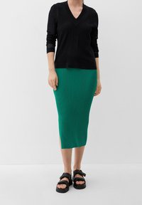 Schwarzer Strickpullover mit V-Ausschnitt, kombiniert mit einem grünen, gerippten Midirock mit Seitenschlitzen und schwarzen klobigen Sandalen.