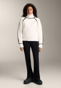 Maglione collo alto bianco e peloso con bordatura nera, abbinato a pantaloni neri a zampa e scarpe platform verde scuro. Design semplice e testurizzato.