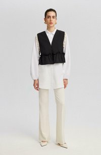 Chaleco negro, cortado con dobladillo con volantes y acentos en color crema, superpuesto sobre una camisa blanca de botones y combinado con pantalones de colores claros.