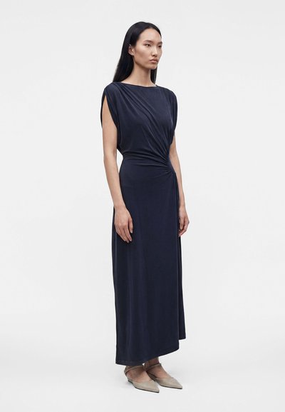 Femme debout portant une robe sans manches bleu marine, longue jusqu'aux chevilles, à taille froncée, et des ballerines beiges à bout pointu sur fond uni.