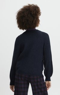 Pull marin à texture, avec un col rond et des manches longues. Porté sur un pantalon à carreaux avec des accents violets.