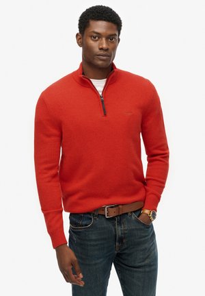 Pullover rosso con colletto a zip, realizzato in materiale morbido. Indossato con jeans blu e una cintura marrone, con un orologio da polso al polso sinistro.