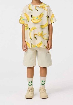 Light beige short-sleeve T-shirt with a banana print, paired with light beige shorts and cream sneakers. Socks feature green smiley faces.