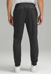 Schwarze Sporthosen mit glatter Textur, elastischem Bund und schmal zulaufenden Beinen. Verfügt über eine Gesäßtasche und Bündchen an den Knöcheln. Weiße Sneakers sind sichtbar.