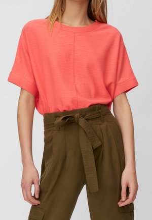 Woman wearing coral short-sleeve top tucked into high-waisted olive green pants with a tied fabric belt and side cargo pockets.