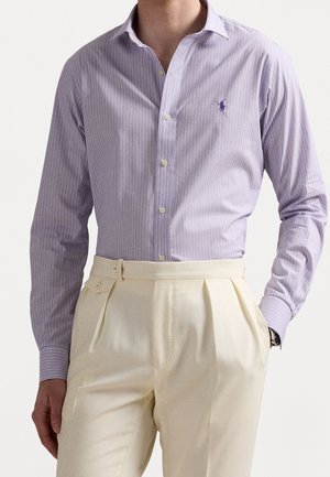 Homme portant une chemise à fines rayures violet clair boutonnée rentrée dans un pantalon crème plissé taille haute avec poches latérales.