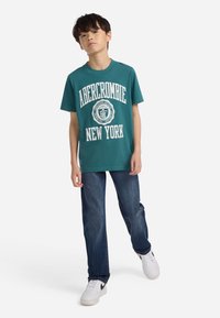 Αγόρι που φοράει μπλουζάκι Abercrombie New York σε τυρκουάζ χρώμα, μπλε τζιν και λευκά αθλητικά παπούτσια, στέκεται με το ένα πόδι ελαφρώς μπροστά σε άσπρο φόντο.