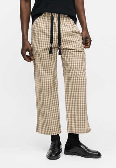 Personne portant un pantalon large à carreaux beige avec un cordon noir, des chaussettes noires, des mocassins noirs et un haut noir sur un fond clair uni.
