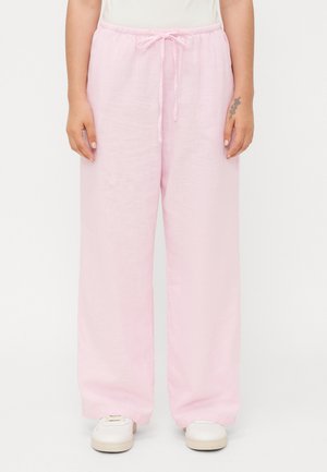 Pantalons larges en lin rose clair avec une taille élastique et un cordon de serrage. Le tissu a une finition légèrement texturée. Portés avec des baskets blanches.