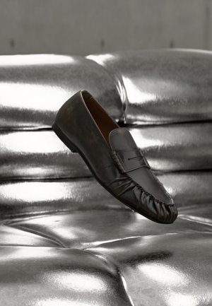 Ένα μονό μαύρο δερμάτινο loafer παπούτσι ακουμπά σε ένα γυαλιστερό μεταλλικό ασημί μαξιλάρι από δέρμα με κουμπιά.