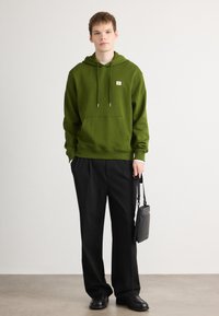 Hanorac verde din bumbac cu buzunar frontal tip kangaroo, șnururi și o mică etichetă cu logo, asortat cu pantaloni negri și o geantă neagră.