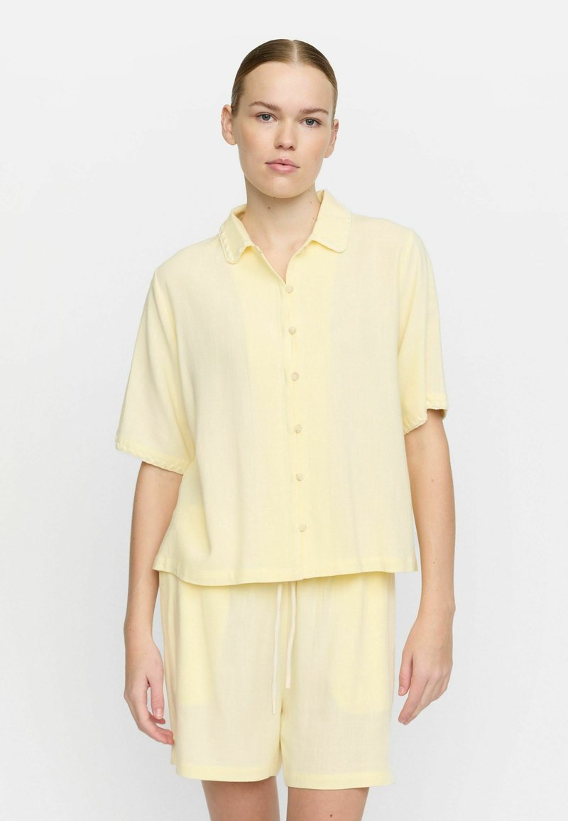 Camicia a maniche corte con colletto e bottoni, abbinata a pantaloni corti in tessuto giallo chiaro, con una consistenza morbida e vita regolabile con cordino.