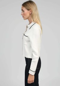 Veste blanche courte en tissu texturé, dotée d'une fermeture à boutons, d'une bordure festonnée noire le long du col et des poignets, ainsi que de poches à l'avant.