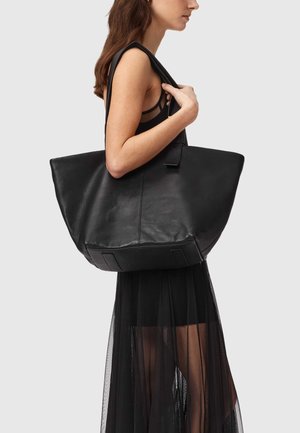 Nainen kantaa suurta mustaa nahkaista olkalaukkua, pukeutuneena hihattomaan mustaan mekkoon, jossa on läpikuultavia helmapaneeleja.