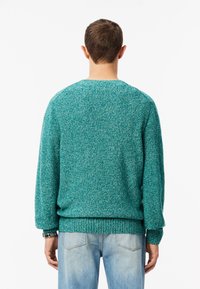 Maglione lavorato a maglia color teal con finitura texturizzata, dotato di scollatura rotonda e polsini e orlo a coste. Indossato sopra jeans azzurri chiari.