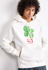 Weißer Hoodie mit einem Grafikdesign von grünen Blättern über einem roten Nike-Logo, mit einer vorderen Kängurutasche und verstellbarem Kapuzenzug. Weicher Stoff.