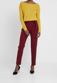 Un pull jaune tricoté à épaules dénudées associé à un pantalon taille haute bordeaux avec une ceinture nouée, complété par des chaussures à talons hauts noires.