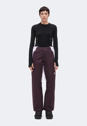 Personne debout, face à l’avant, portant un haut noir à manches longues et un pantalon de ski violet foncé avec des bretelles ainsi que des bottes noires sur un fond clair.
