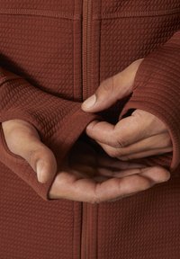 Chaqueta texturizada de color óxido con cierre de cremallera, que cuenta con agujeros para los pulgares en las mangas y detalles de costura visibles. Las manos están colocadas cerca de la cintura.