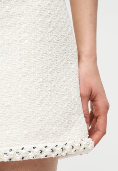 Gros plan sur une jupe blanche texturée ornée de perles et de strass le long de l'ourlet, accompagnée de la main d'une personne reposant à côté.