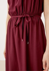 Vestido burdeos con escote redondeado, cintura elástica y detalle de lazo de tela. Textura suave con un ligero brillo, con acentos plateados.