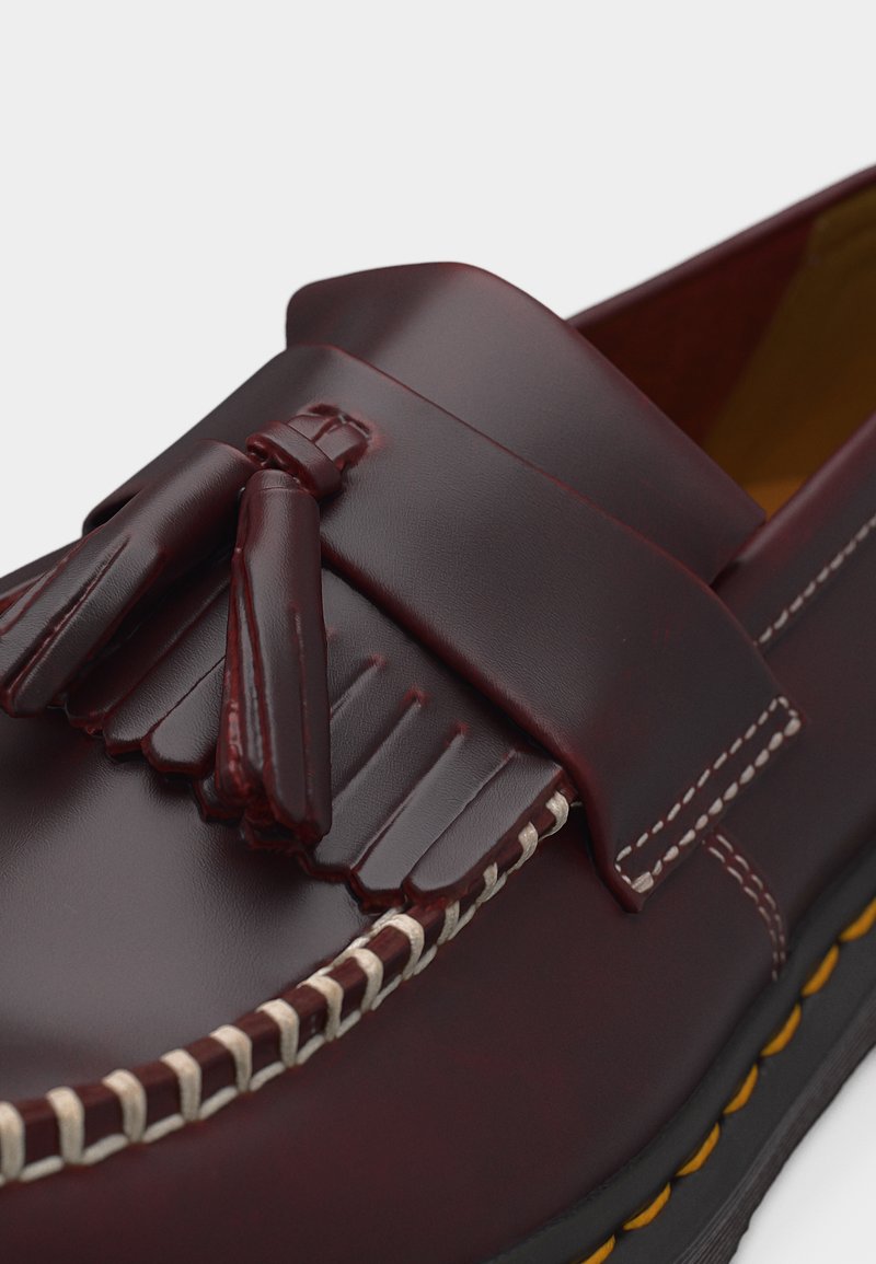 Mocassino in pelle bordeaux scuro con nappine, cuciture bianche, dettaglio a frange e suola nera con cuciture gialle, primo piano.