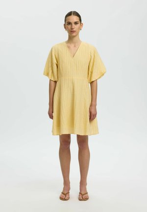 Kvinde iført en kort, tekstureret gul kjole med pufærmer og V-hals, der står foran en ensfarvet lys grå baggrund.