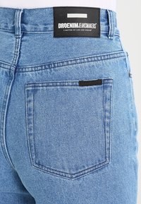 Světle modré džíny z denimu s černou etiketou a bílým textem na pase, tradiční design kapes a hladká textura.