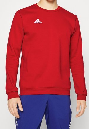 Mann trägt einen roten Adidas-Sweatshirt und blaue Adidas-Hose, gezeigt vom Hals bis zur Mitte des Oberschenkels mit entspannten Armen an den Seiten.