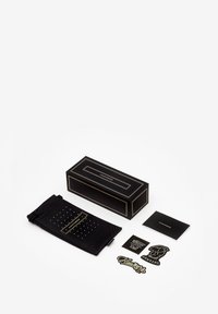 Mustaline musta ristkülikukujuline karp kuldsete aktsentidega, must kott täppidega ning mitmed kleebised koos info kaardiga.