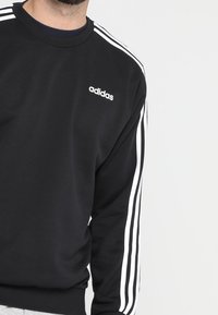 Sweat-shirt noir avec logo Adidas blanc sur la poitrine. Présente des bandes blanches sur les deux manches. Fabriqué dans un tissu doux et texturé.