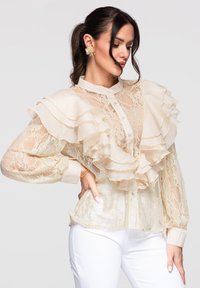 Femme portant un chemisier en dentelle crème transparente avec de grands volants et des boutons en perles, associé à un pantalon blanc, posant avec une main sur la hanche.