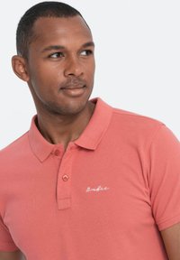 Korallinvärinen polo-paita, joka on valmistettu puuvillasta, klassisella kauluksella, kaksikappaleisella napitusosalla ja brodeeratulla logolla rinnassa.