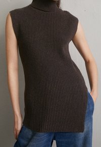 Rippstrick, ärmelloser Rollkragenpullover aus dunkelbraunem Strickstoff mit seitlichen Schlitzen und taillierter Silhouette. Getragen mit hochgeschnittenen blauen Jeans.