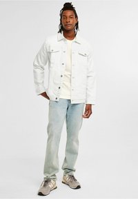 Weiße Jeansjacke mit vorderen Taschen, kombiniert mit einem beigen T-Shirt und hellblauen Jeans, getragen mit grauen und beige Sneakers.