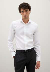 Uomo che indossa una camicia bianca ben stirata infilata in pantaloni con righe nere scure, con una cintura nera, in piedi contro uno sfondo chiaro e neutro.