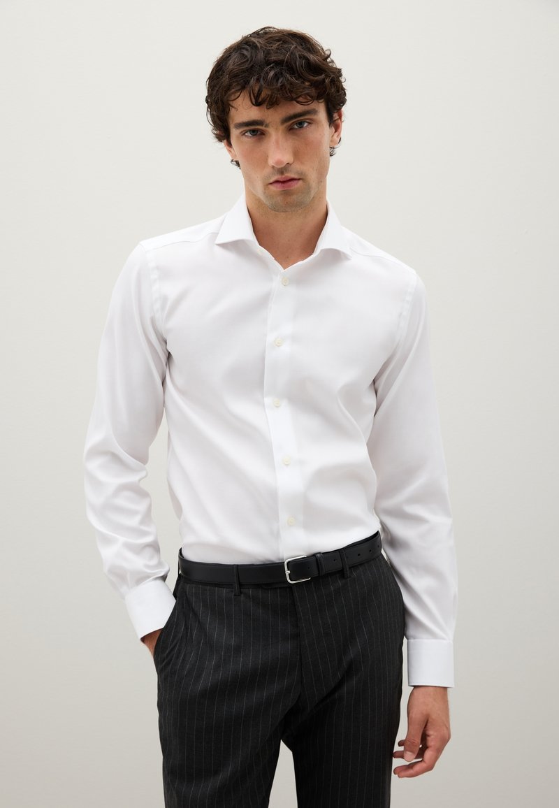 Uomo che indossa una camicia bianca ben stirata infilata in pantaloni con righe nere scure, con una cintura nera, in piedi contro uno sfondo chiaro e neutro.