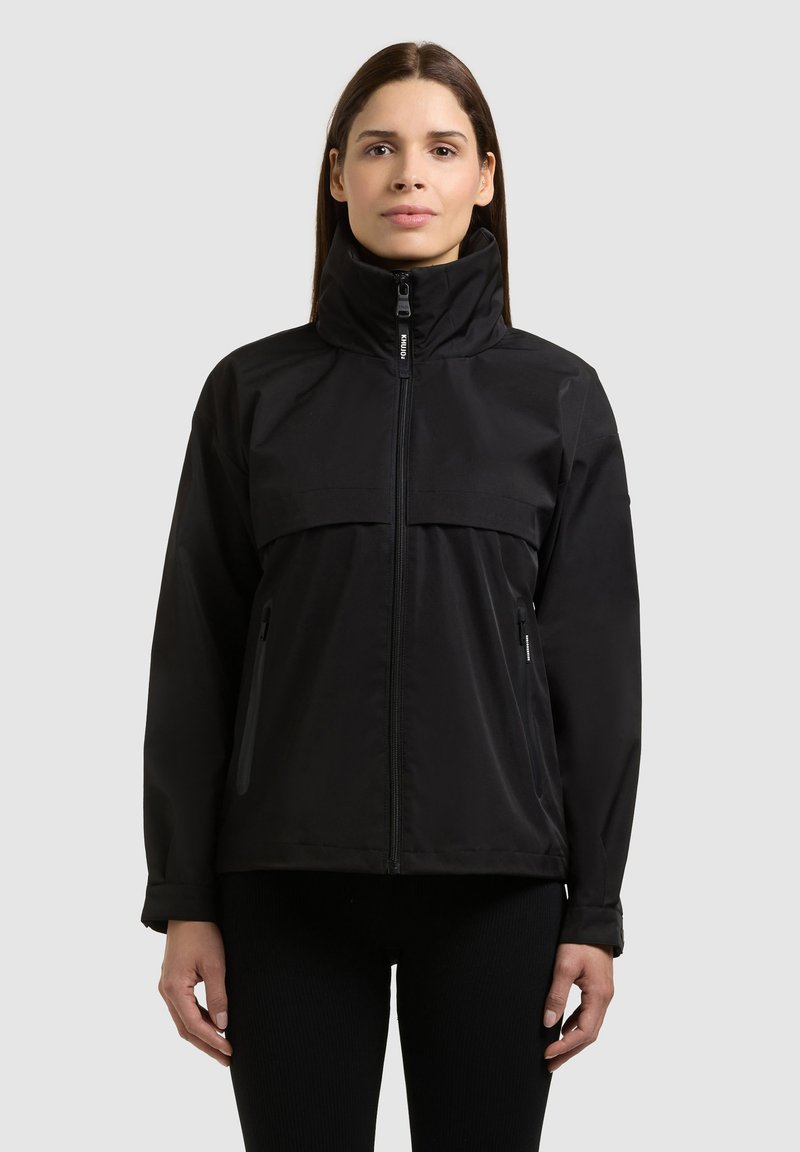 Femme portant une veste noire à fermeture éclair avec col montant et poches zippées latérales, debout devant un fond gris clair uni.
