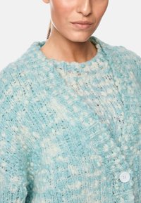 Blau gestrickte Strickjacke mit einem strukturierten Muster, das verschiedene Blautöne und Weiß umfasst, einem runden Ausschnitt und einem weißen Knopfdetail.