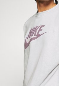 Sudadera gris claro de tejido suave con un destacado logo púrpura de Nike, en un diseño deportivo, con cuello redondo y mangas largas.