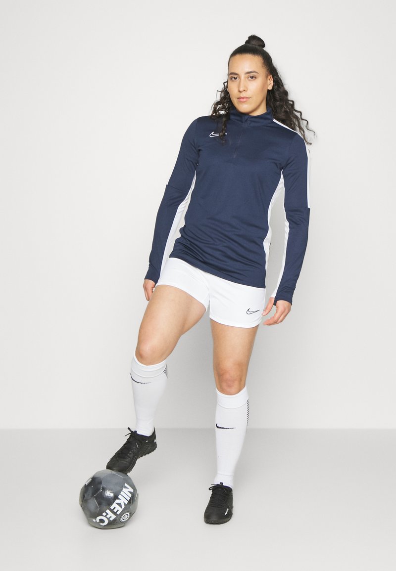 Marineblaues langärmeliges Sportoberteil mit weißen Seitenpaneelen, kombiniert mit weißen Shorts und Socken, steht neben einem schwarz-grauen Fußball.