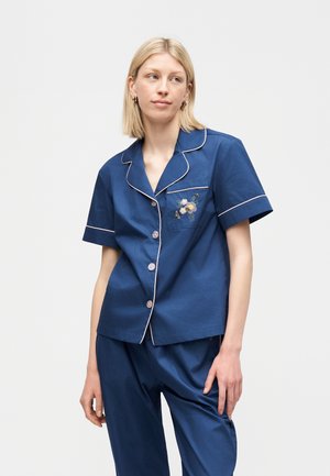 Γυναίκα που φοράει σετ πυτζάμες μεσίστιου μπλε χρώματος, κοντομάνικο, με λευκή διακόσμηση και κεντήματα με λουλούδια στην τσέπη στο στήθος, στέκεται μπροστά σε απλό φόντο.