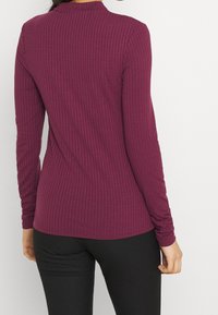 Langärmliges Oberteil aus dunkelviolettem Rippstoff mit hohem Kragen und taillierter Passform, kombiniert mit schwarzen Slim-Fit-Hosen.