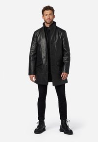 Black leather coat with a collar, front placket buttons, and two front pockets. Worn over a black outfit and paired with black boots.