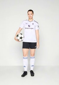 Jogadora de futebol jovem, de pé, segurando uma bola com padrão de estrelas em preto e branco, vestindo uma camisola branca, calções pretos e meias brancas até ao joelho.