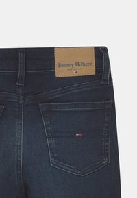Vaqueros de mezclilla azul oscuro con una etiqueta de cuero marrón que dice "Tommy Hilfiger" y un pequeño logo en rojo, blanco y azul en el bolsillo trasero.